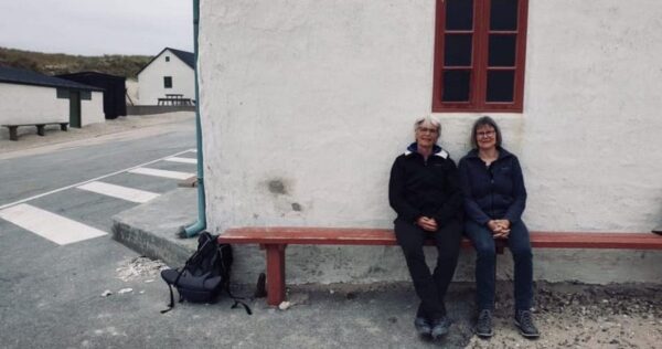 To kvinder sidder på en råd bænk foran et hvidt hus tæt på stranden