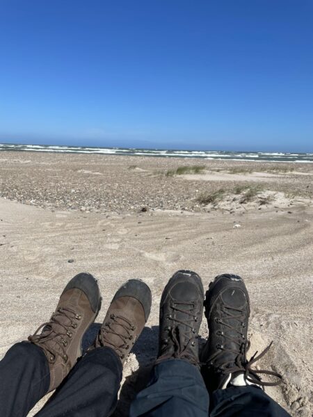 To personer sidder i sandet ved stranden, hvor man kan se deres vandrestøvler og havet i horisonten