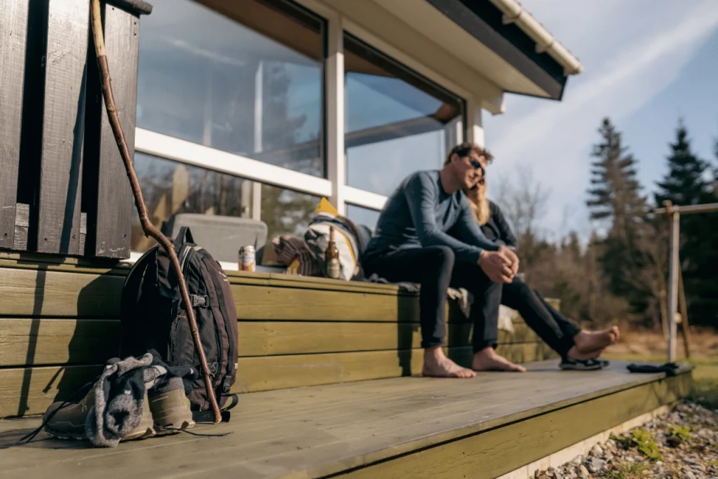 Par nyder et hvil i solen på sommerhusets terrasse- Rygsæk, vandrestav og vandrestøvler står i forgrunden af billedet.