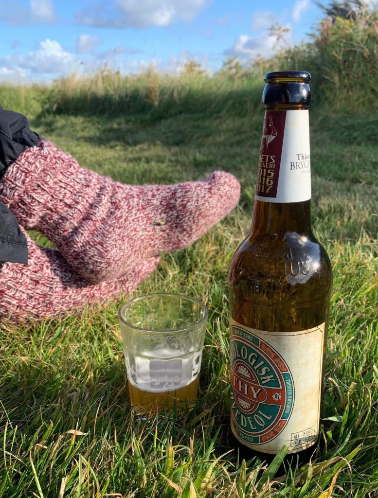 Økologisk hvedeøl på flaske fra Thisted Bryghus står i græsset på en solskinsdag lidt af øllen er hældt op i et glas som står ved siden af en persons fødder iklædt et par røde uldsokker er i baggrunden