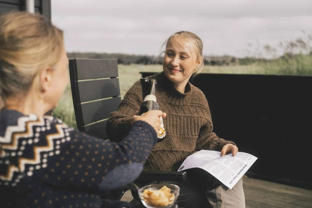 To kvinder skkåler i flaskeøl på terrassen ved sommerhuset. På bordet står en skål med chips og den ene kvinde sidder med Oh So Quiets infomatriale